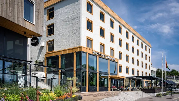 Quality Hotel Grand Larvik, en modern byggnad med vit fasad och gyllene detaljer, omgiven av livfulla blommor under en blå himmel.