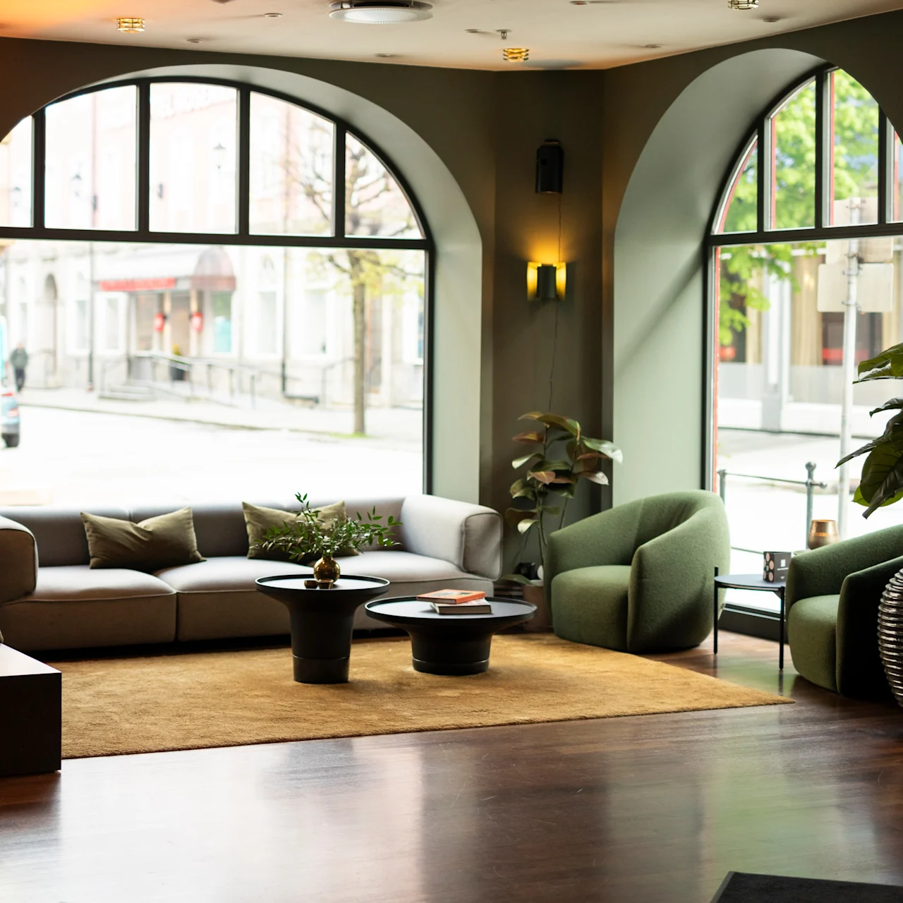 En moderne lounge har en sofa og lænestole omkring to små borde på et tæppe, oplyst af store buede vinduer med udsigt til gaden. Potteplanter tilføjer grønne nuancer.