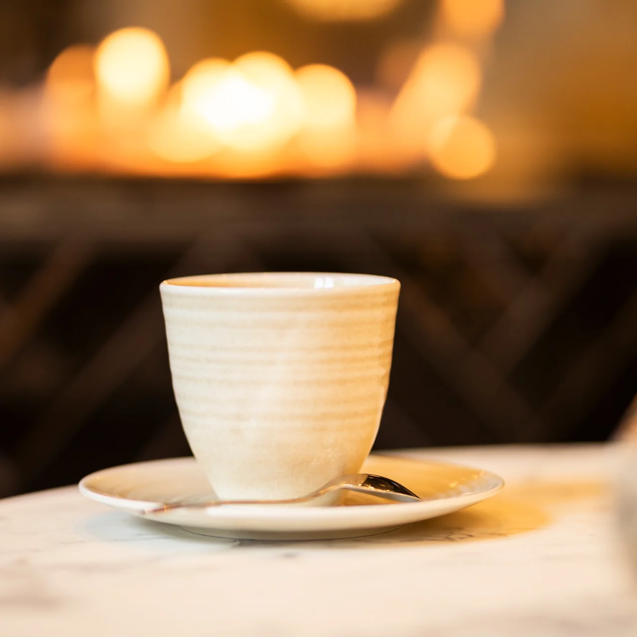 A ceramic cup rests on a saucer with a spoon, placed on a marble table. Warm, blurred lights glow softly in the background, suggesting a cozy setting.