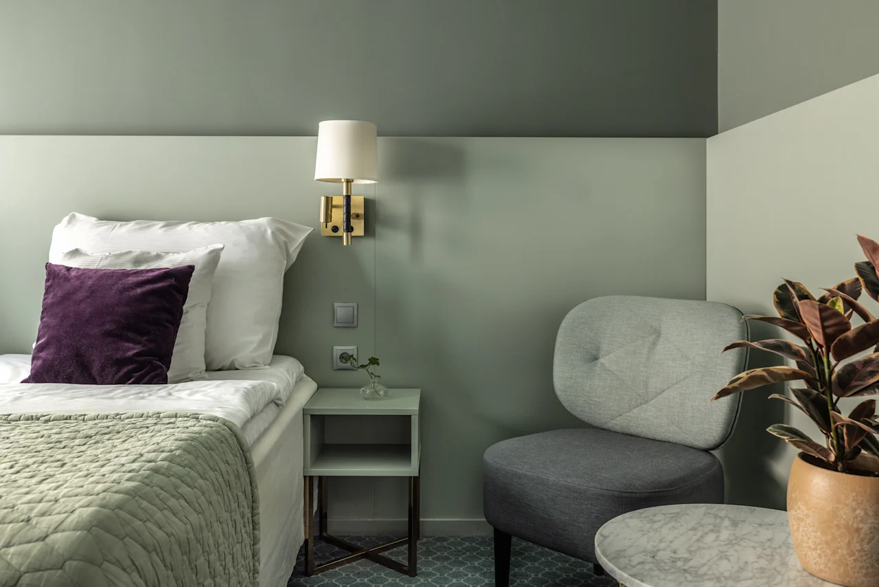 Miellyttävä makuuhuoneen nurkkaus, jossa on siististi pedattu sänky, pehmeät tyynyt, pieni yöpöytä, mukava nojatuoli ja koristekasvi tuomassa luontoa. Seinälamppu valaisee rauhallisen näkymän.