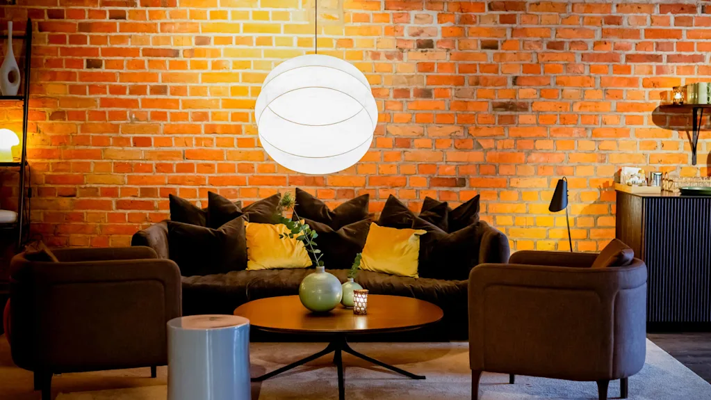 A cozy lounge area with a sofa, armchairs, and a coffee table set against an exposed brick wall, lit by a stylish pendant light.
