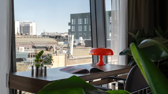 Clarion Hotel Sign - Stratum Suite. A stylish desk with a red lamp and magazine, overlooking a city skyline from a large window.