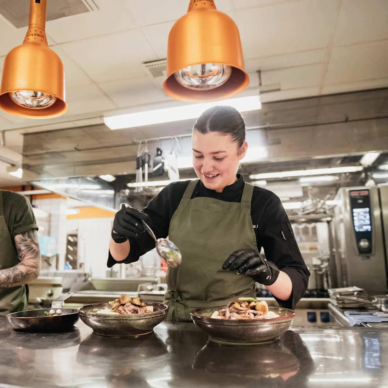 Two chefs in green aprons prepare dishes in a professional kitchen. One garnishes a plate, while the other ladles food into a bowl. Copper pendant lights illuminate the workspace.