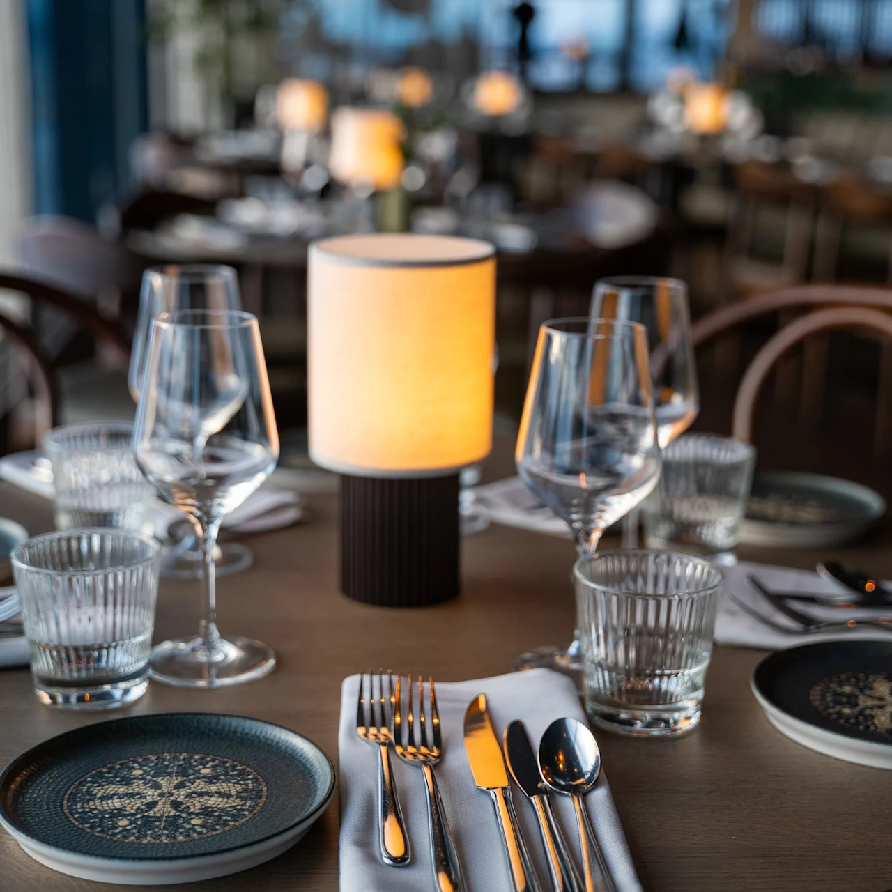 Table elegantly set with plates, wine glasses, and cutlery on a white napkin; a small lamp provides warm light. Soft, hanging lights create a cozy restaurant atmosphere.