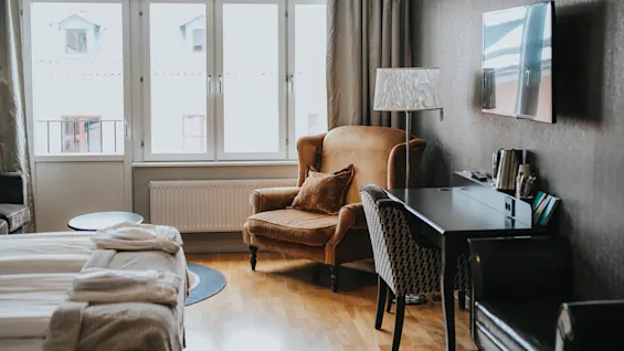 Et hyggeligt værelse med en vintage lænestol ved et vindue, en seng med tæppe, et skrivebord med bøger og stemningsfuld belysning, der skaber en varm og indbydende atmosfære.