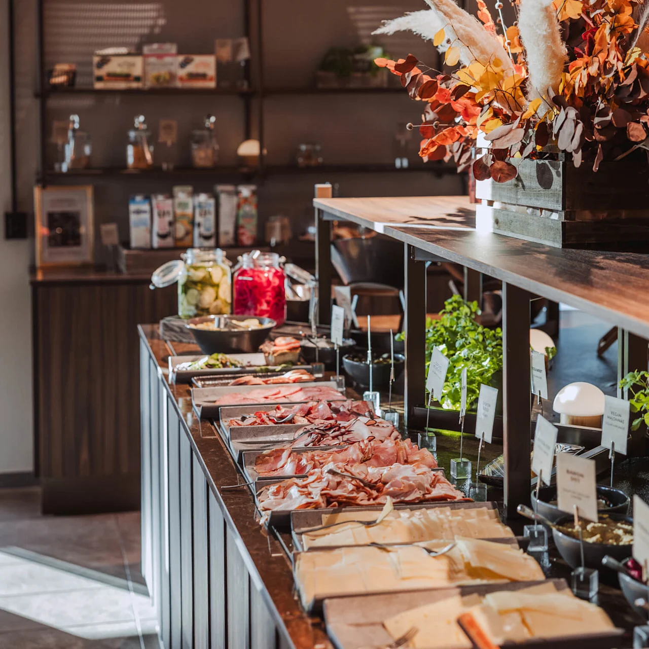 Ett buffébord visar olika charkuterier och ostar, medan livfulla blomarrangemang och en varm taklampa skapar en inbjudande matsalsmiljö. Hyllor med olika föremål är synliga i bakgrunden.
