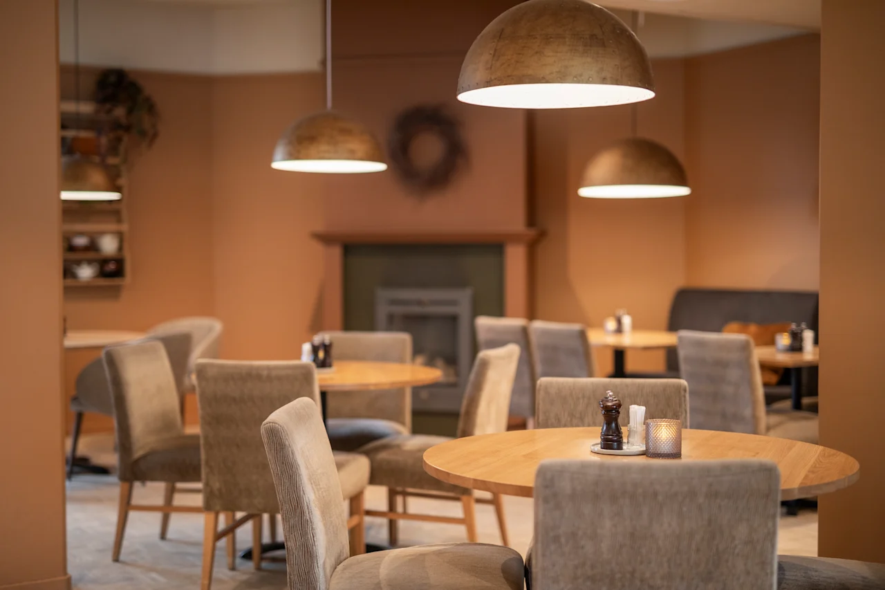 Tables and chairs arranged in a cozy cafe setting, illuminated by dome-shaped pendant lights. The warm, brown walls create an inviting ambiance with a decorative wreath above a fireplace.