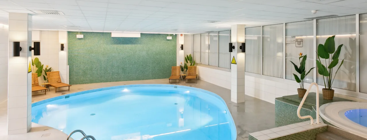 Swimming pool–calm water in brightly lit indoor setting, surrounded by lounge chairs and potted plants; adjacent whirlpool on tiled platform.