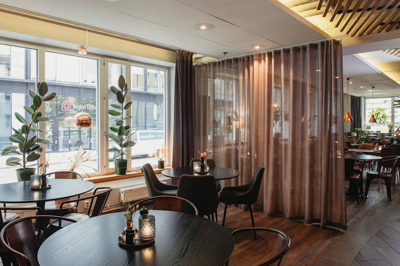 Home Hotel Uppsala restaurant with dark tables, chairs, and large windows. Outside, a street with a Region Upps sign. Sheer curtains.