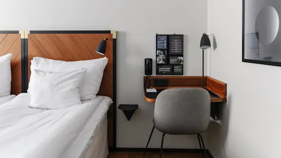 En snyggt bäddad säng med vitt lakan bredvid ett trä nattduksbord med en telefon, en lampa och ett informationskort i ett prydligt hotellrum.