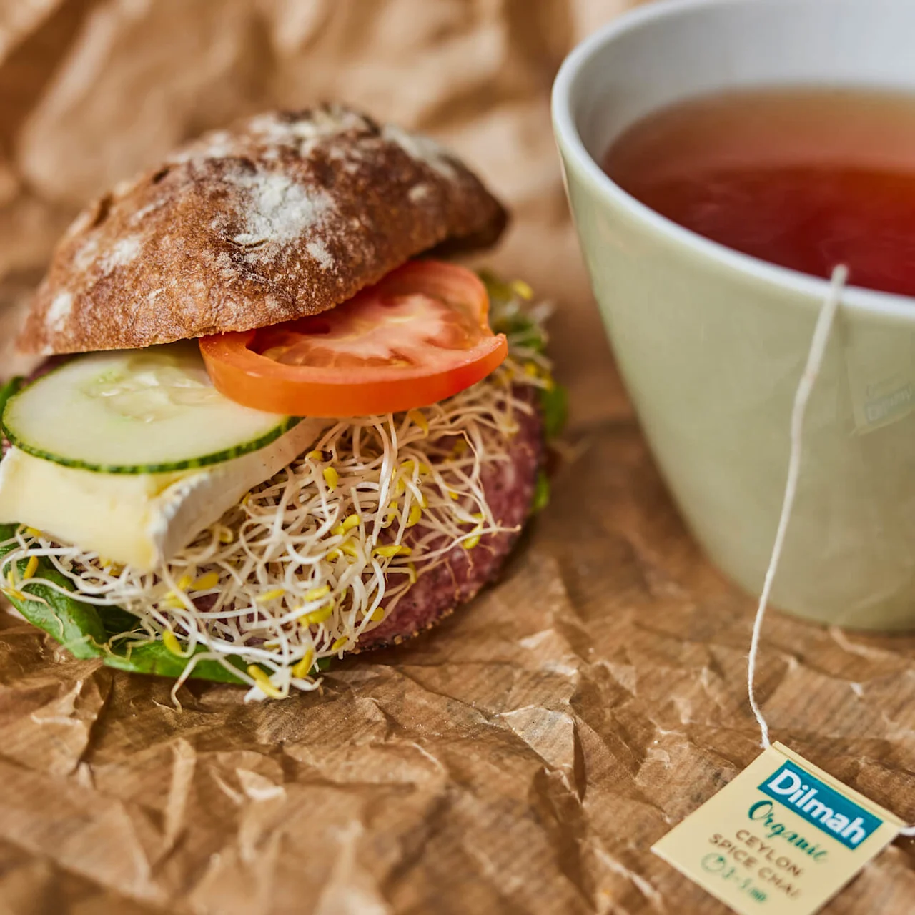 En sandwich med salat, spirer, ost, agurk og tomat ligger på krøllet brunt papir ved siden av en kopp te. Teetiketten sier: "Dilmah Organic Ceylon Spicy Chai."