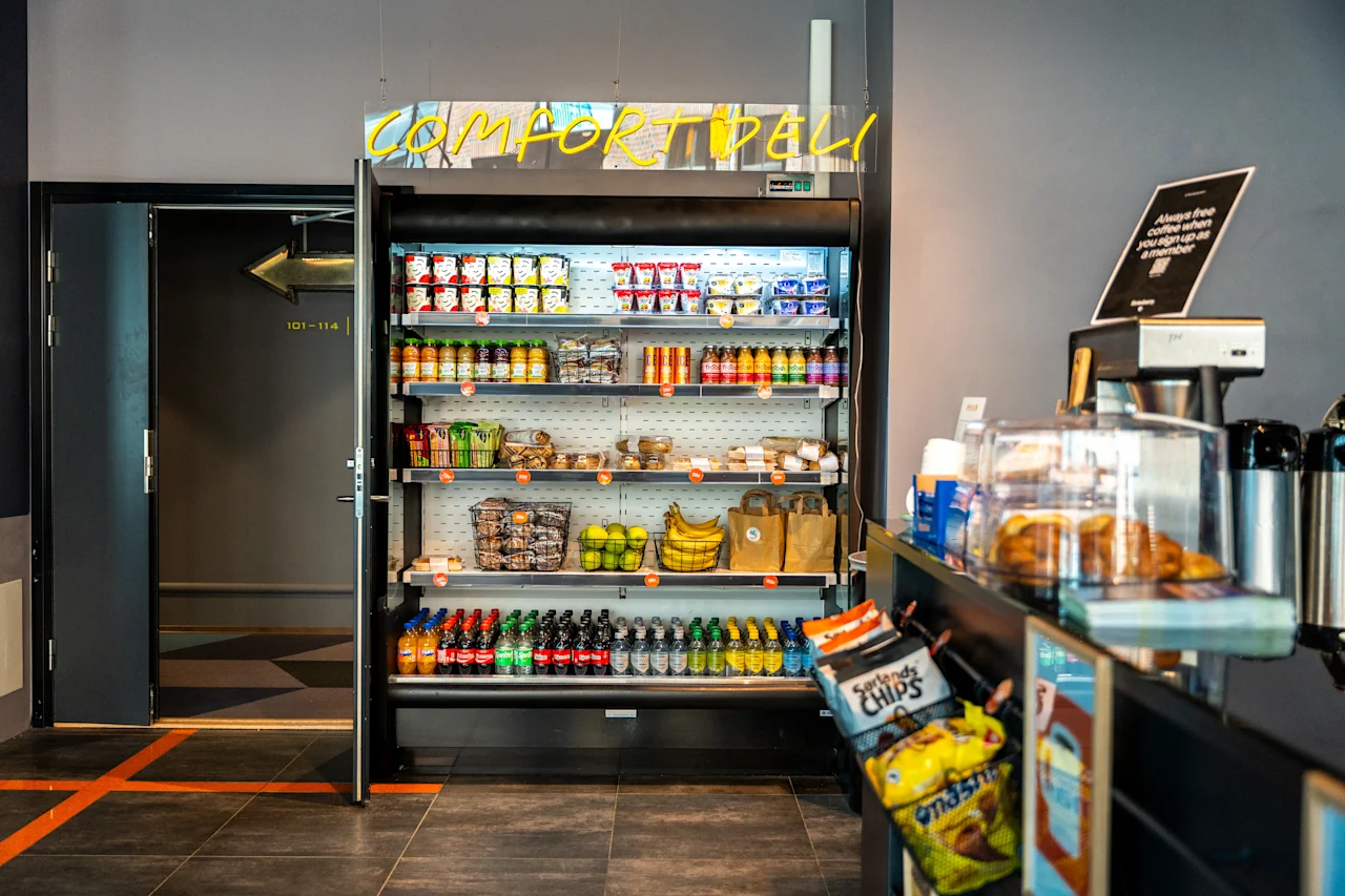 Et køleskab indeholder snacks, drikkevarer og frugt. Ovenover ses et neonskilt med teksten "COMFORT DELI". I nærheden er der en disk med chips og bagværk. En dør mærket "403-416" fører til en gang.