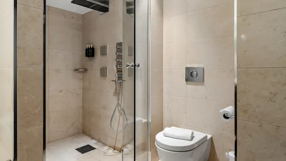 En elegant glashytte med moderne inventar står i et beige-flisebelagt badeværelse, ved siden af et toilet med et foldet hvidt håndklæde ovenpå.