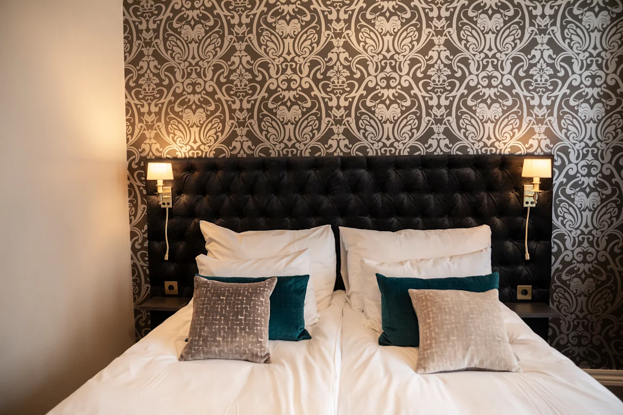 Et hyggeligt hotelværelse med en stor seng, bløde puder og elegant mønstret tapet, oplyst af varme sengelamper.