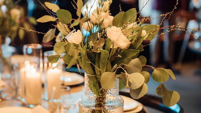 A flower bouquet in a glass vase sits on a wooden dining table set with plates, candles, and cutlery. People gather in the softly lit background.