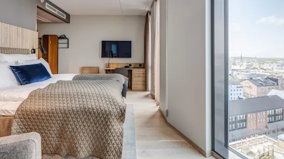 En pent seng med grått dyne står mot en veggmontert TV. Store vinduer gir panoramautsikt over byen og fyller det moderne rommet med naturlig lys.