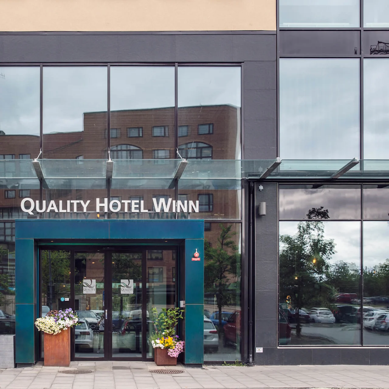 Quality Hotel Winn-entré med automatiska glasdörrar, flankeras av blomarrangemang i en modern stadsmiljö, med reflektioner av närliggande byggnader.
