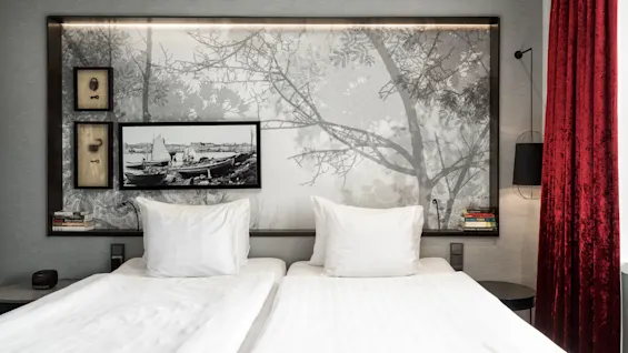Two neatly made beds with white linens, framed black and white artwork above, in a modern bedroom with a red curtain.
