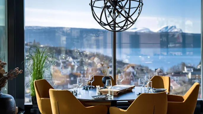 A dining table set with glasses and plates is surrounded by mustard-yellow chairs. The setting is near large windows with a scenic view of mountains and a town.