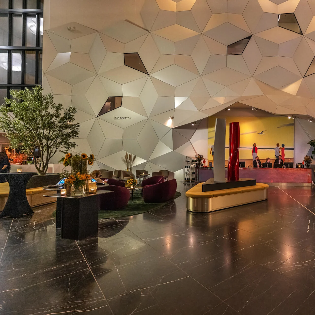 A geometric wall with hexagonal patterns overlooks a spacious lobby with modern furniture and artwork. Text on the wall reads "THE ROOFTOP." Plants and seating areas are present.