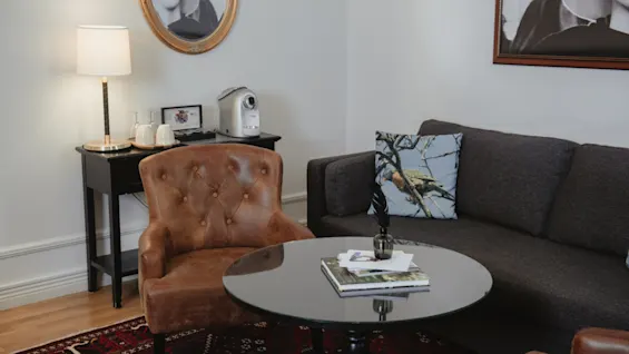 En brun lænestol og grå sofa omgiver et glasbord med bøger i en hyggelig stue med tæppe, vægkunst og lampe på et sidebord med kaffemaskine.