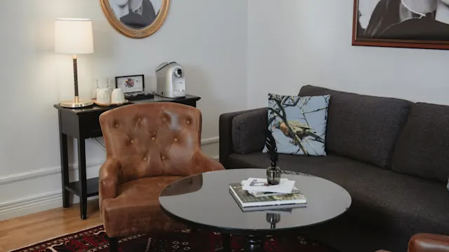 En brun fåtölj och grå soffa omger ett glasbord med böcker i ett mysigt vardagsrum med matta, väggkonst och lampa på ett sidobord med kaffemaskin.