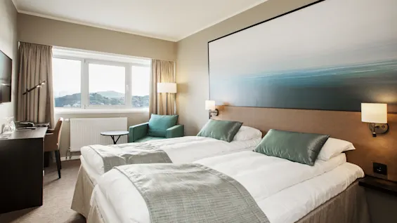 Et hotellrom med to oppredde senger, en komfortabel stol og et skrivebord ved et vindu. Vinduet gir utsikt over fjerne åser.