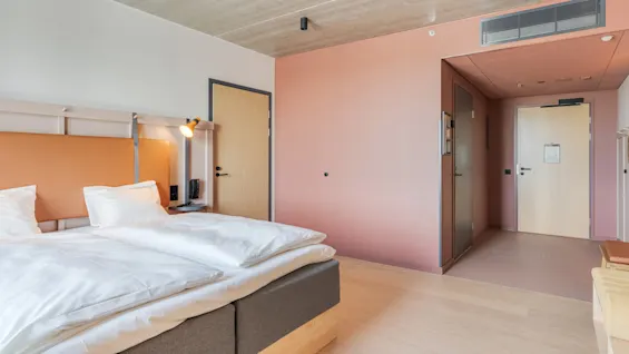 En pæn seng med hvidt sengelinned og en moderne lampe på væggen. Beliggende i et minimalistisk rum med lyst træ og lyserøde vægge, er to lukkede døre synlige.