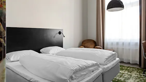 Two neatly made single beds in a small room with a black headboard and adjustable reading lamps. A brown chair sits by the window, beside long beige curtains.