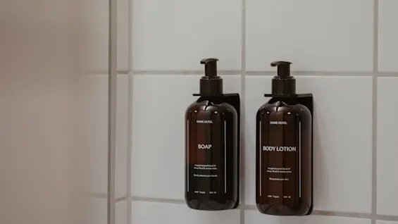 Kaksi tummanruskeaa pumppupulloa, toinen SAIPPUA ja toinen BODY LOTION, valkoisella kaakeliseinällä.