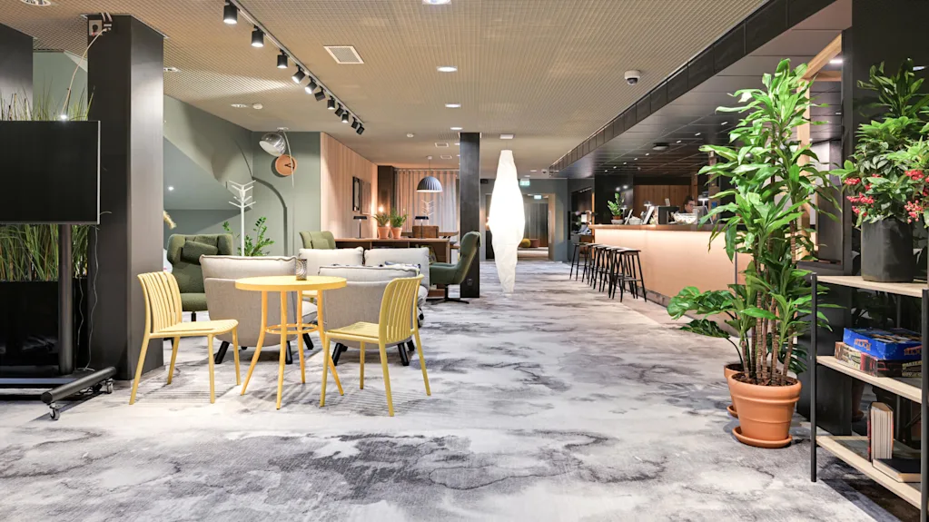 Et loungeområde med gule stoler og et bord foran, omgitt av sofaer og planter. En bardisk med krakker er til høyre.