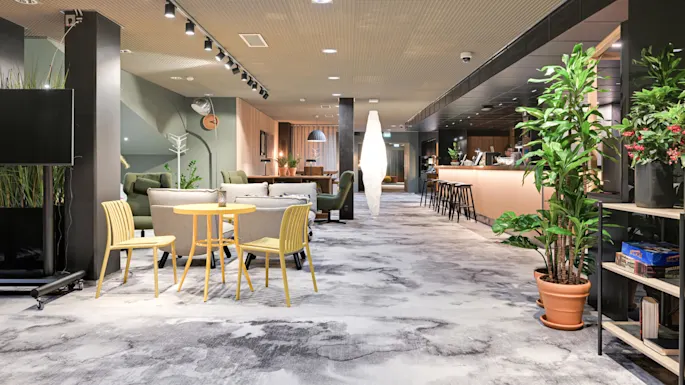 Et loungeområde med gule stoler og et bord foran, omgitt av sofaer og planter. En bardisk med krakker er til høyre.