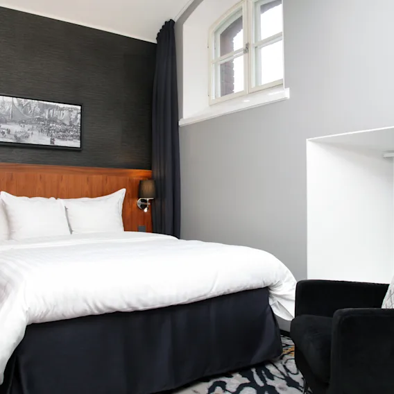 Viihtyisä hotellihuone, jossa on mukava sänky, tumma nojatuoli ja kehystettyä taidetta seinällä, tarjoten rentouttavan oleskelun.