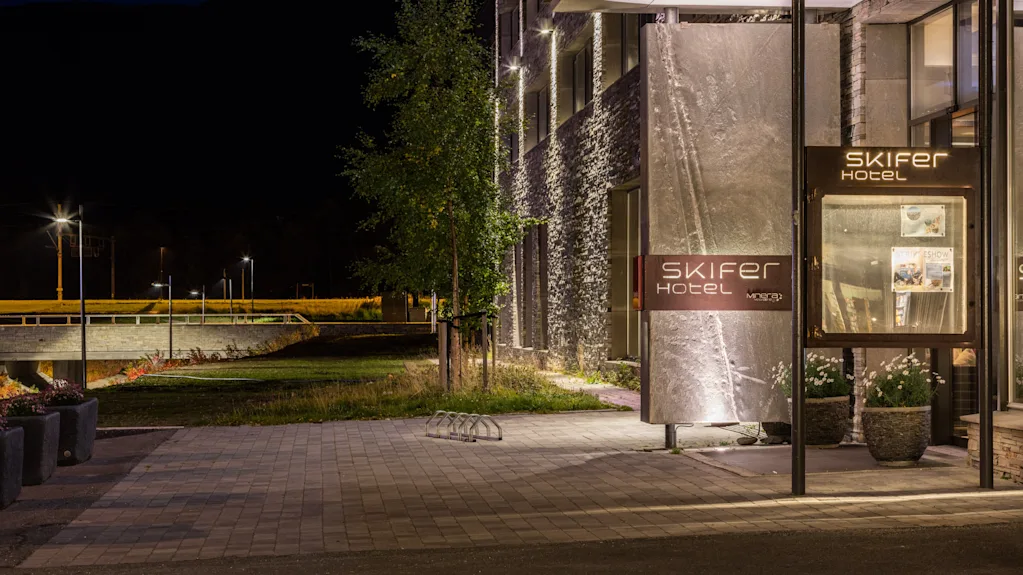 En skylt lyder "SKIFER HOTEL" vid en upplyst entré till en modern byggnad på natten, omgiven av träd och en kullerstensväg. I närheten syns en bäck med en avlägsen bro.