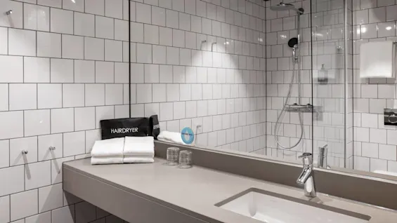 A modern bathroom showcases a countertop with a white sink, folded towels, a hairdryer bag labeled "HAIRDRYER," glasses, and tiled walls; a shower is enclosed in glass.