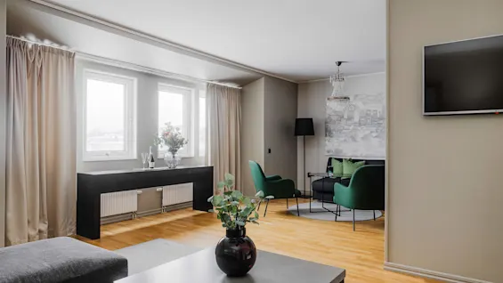 En vas med blad står på ett bord i ett modernt vardagsrum. Gröna stolar omger ett bord, med en ljuskrona ovanför och en TV på väggen.