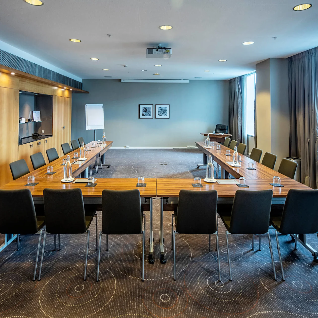 Konferensbord dukat med vattenflaskor och glas i ett rymligt, väl belyst mötesrum med träpaneler och stora fönster med gardiner. Ett blädderblock och konstverk dekorerar väggarna.