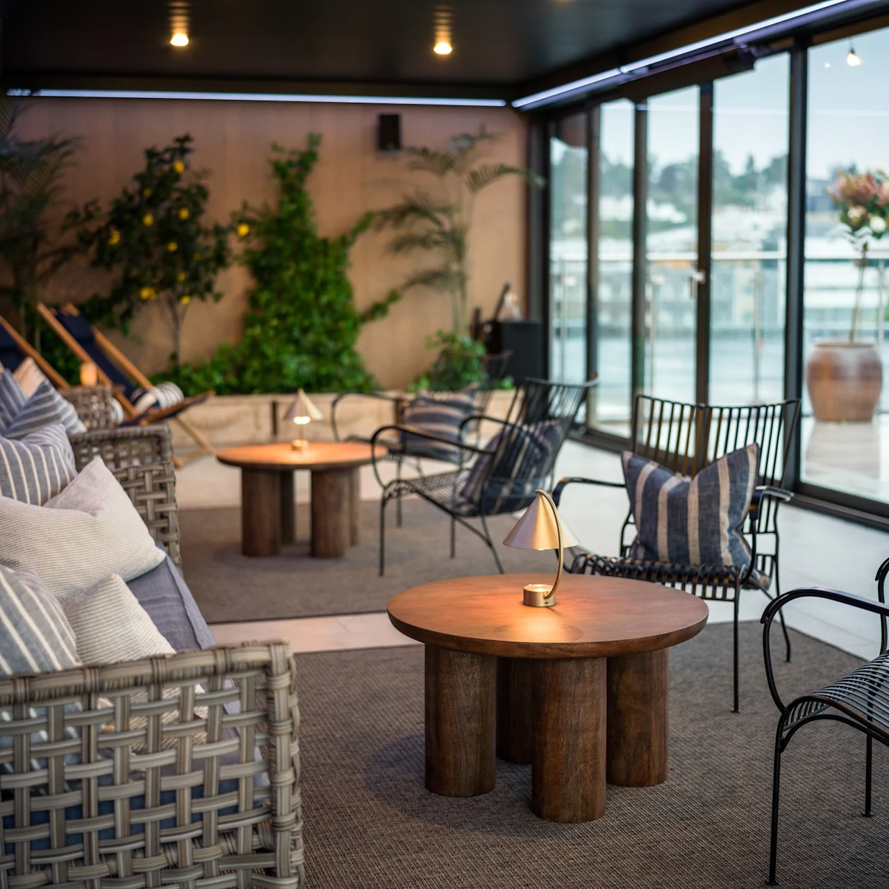 Bekväma sittplatser omger träbord med små lampor i en mysig inomhuslounge med stora fönster och krukväxter som skapar en avslappnad och inbjudande atmosfär.