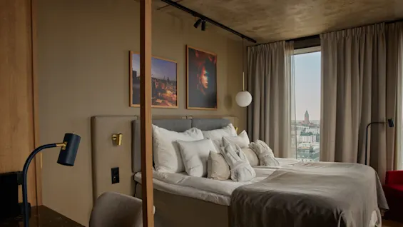 En snyggt bäddad säng med kuddar och morgonrockar står i ett hotellrum, omgiven av två tavlor på väggen. En stadsutsikt syns genom golv-till-tak-gardiner.