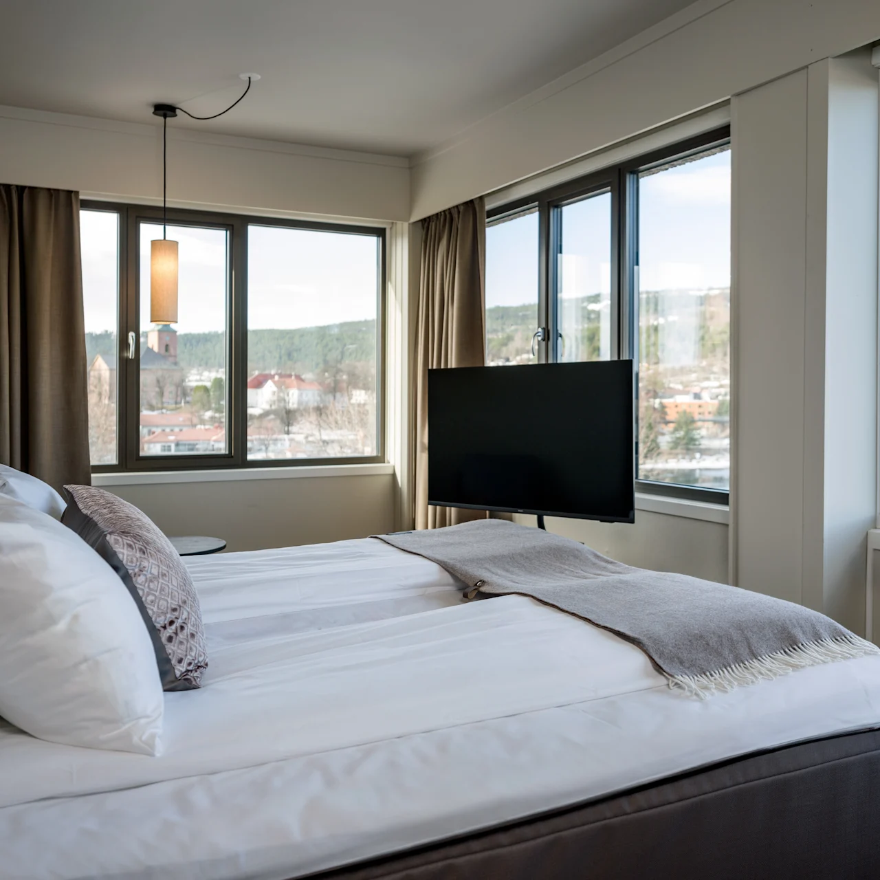 A cozy bedroom features a neatly made bed with white linens. A TV is mounted in front. Large windows provide a view of trees and buildings outside.