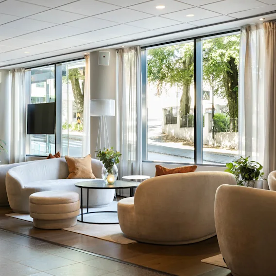 Et moderne loungeområde med hvite sofaer rundt små bord i et solfylt rom med store vinduer mot grønne omgivelser. Ingen tekst er synlig.