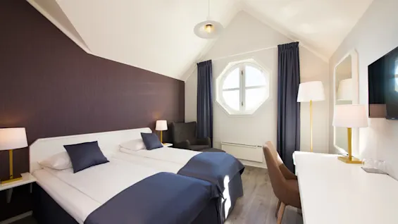 Ett välbelyst hotellrum med två enkelsängar, stolar, bord och ett åttakantigt fönster, som visar en ren, modern design.