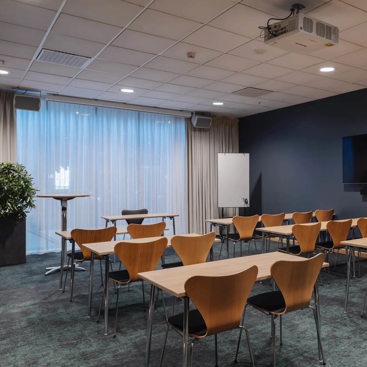 Ett konferensrum fyllt med trästolar och bord vetter mot en stor väggmonterad skärm. Gardiner täcker fönstren, och en projektor är monterad i taket.