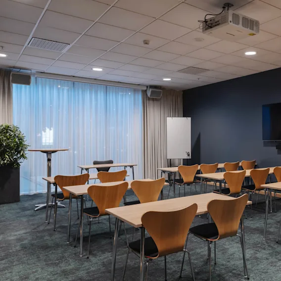 Ett konferensrum fyllt med trästolar och bord vetter mot en stor väggmonterad skärm. Gardiner täcker fönstren, och en projektor är monterad i taket.