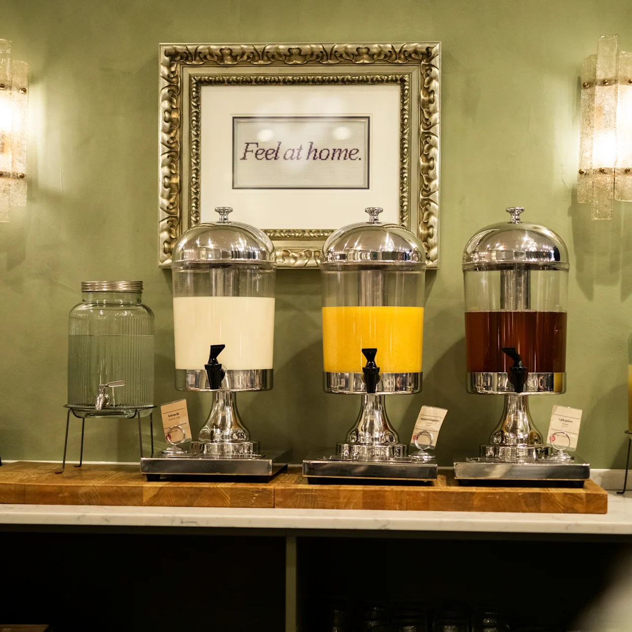 Water dispensers with various drinks stand lined up on a counter. Framed text reads "Feel at home," with soft lighting on a green wall providing a cozy atmosphere.
