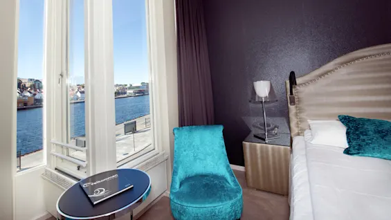 A hotel room with a bed, chair, and a window overlooking water and buildings.