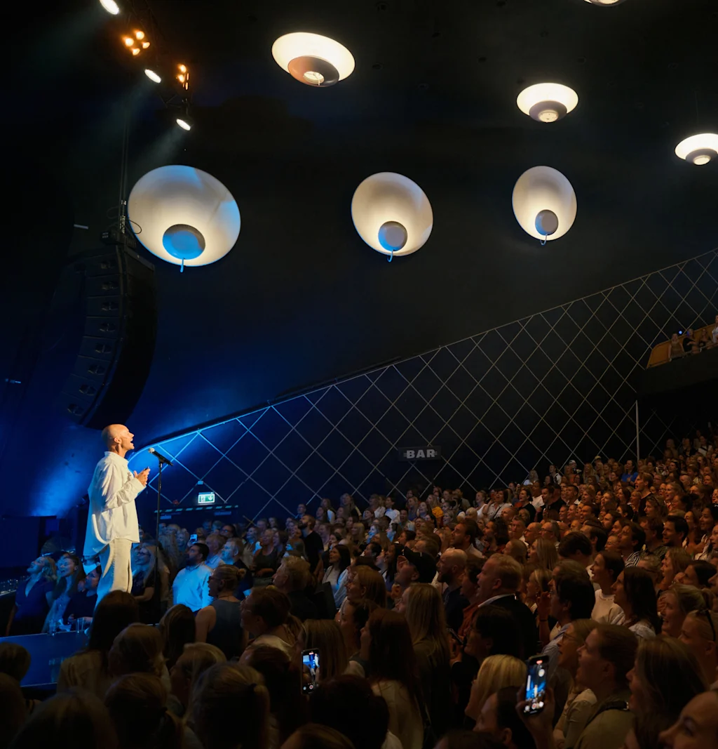 En taler henvender seg til et stort, engasjert publikum i en mørk sal med unike sirkulære taklamper. Et "BAR"-skilt er synlig.