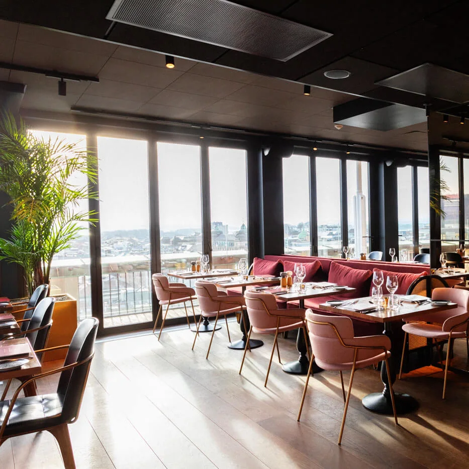 A bright, modern restaurant with wooden floors features pink and black chairs around set tables. Large windows provide a city view, and lush green plants enhance the elegant atmosphere.