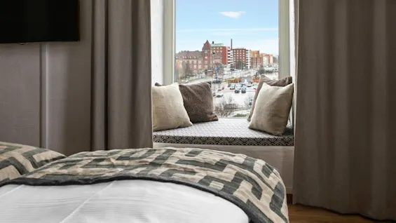 A bed foregrounding a window view of an urban landscape, with patterns and pillows enhancing interior comfort.