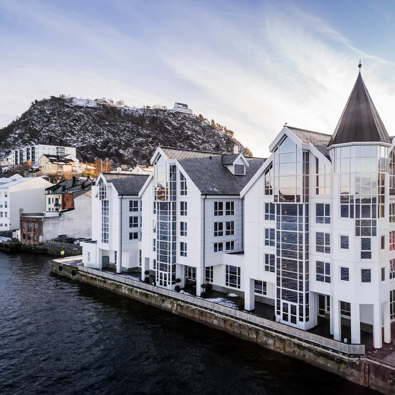 En stor hvid bygning med spidse tage spejler sig i det rolige vand, mod sneklædte bakker og tilstødende bystrukturer under en blå himmel.